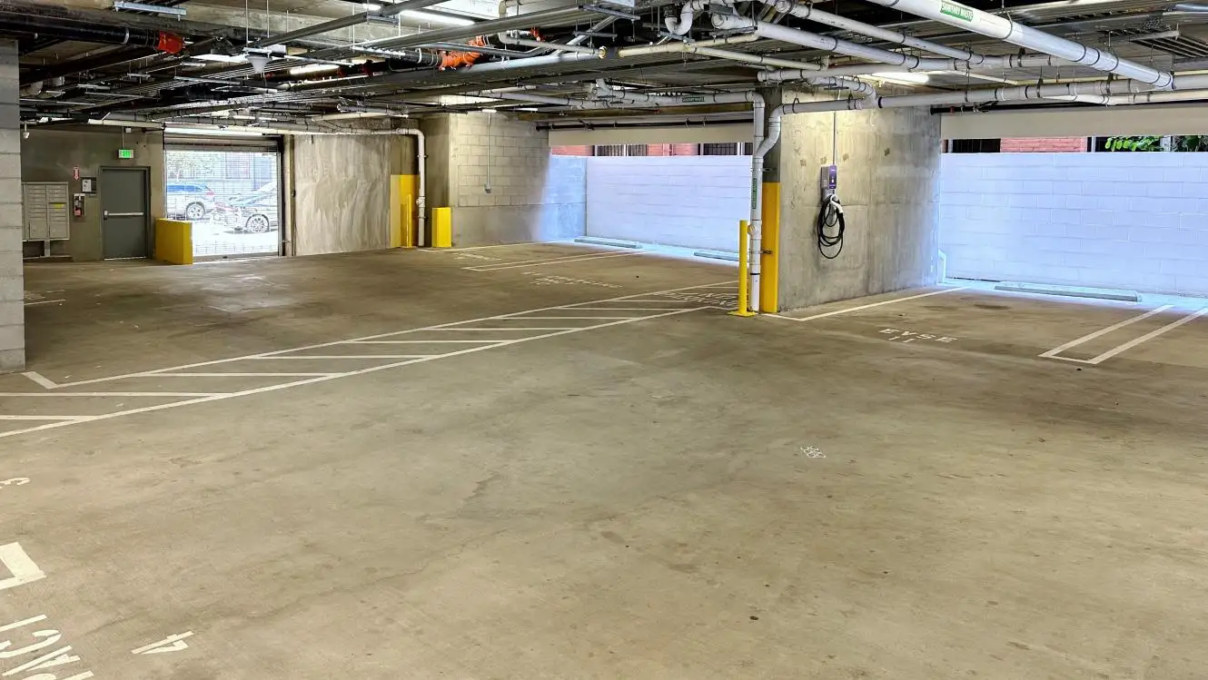 Spacious underground parking garage with marked parking spaces, yellow safety bollards, exposed ceiling piping, and an electric vehicle charging station on the wall.